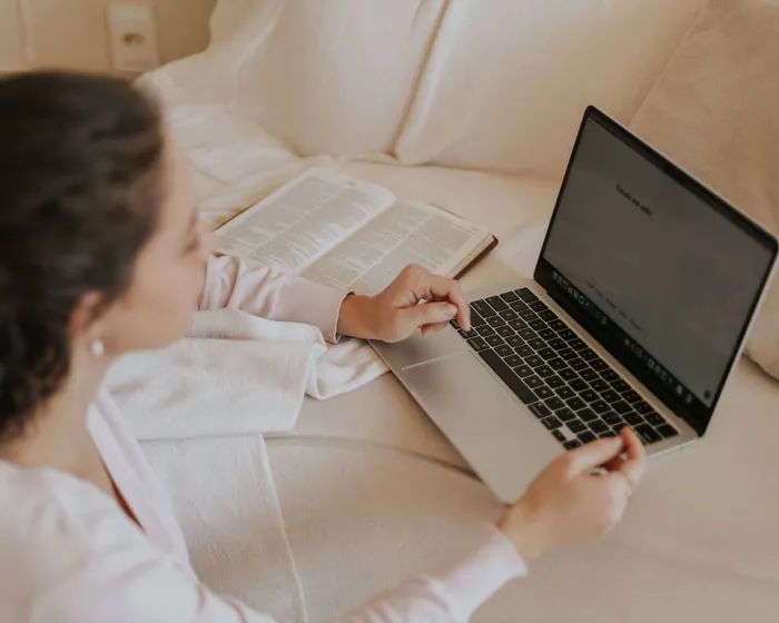 Mulher estudando no notebook no sofá de casa, com um livro aberto ao lado